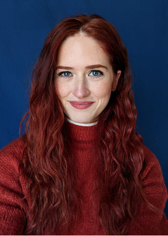 A person with long red hair wearing a burnt orange sweater, against a blue background.
