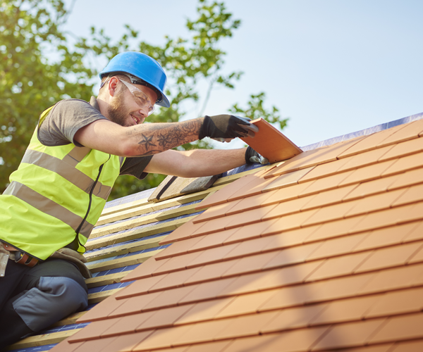 A person wearing a green vest and a blue safety helmet putting a shingle on a roof.