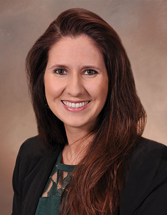 A person with long brown hair wearing a green shirt with a black blazer, against a brown background.