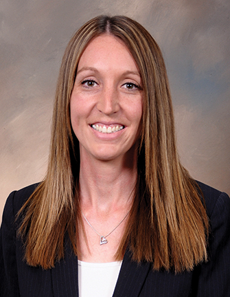 A person with long brown hair wearing a white shirt with a black blazer, against a brown background.