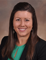 A person with long brown hair wearing a green shirt with a black blazer, against a brown background.