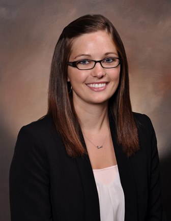 A person with medium brown hair wearing a white shirt with a black blazer, against a brown background.