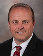 A person with short brown hair wearing a white shirt with a black blazer and a burgundy tie, against a brown background.