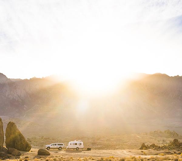 A truck with a travel trailer is parked in front of a mountain with the sun shining over it.