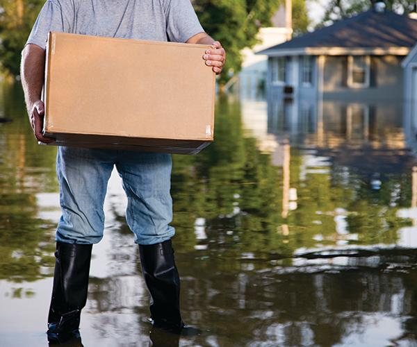 A person wearing rain boots is standing in ankle deep water in front of a house with a box in their hands.