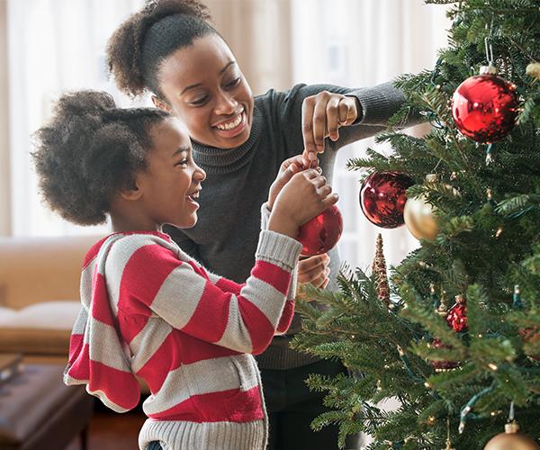 A person and a child are holding a red ornament in front of a decorated tree.