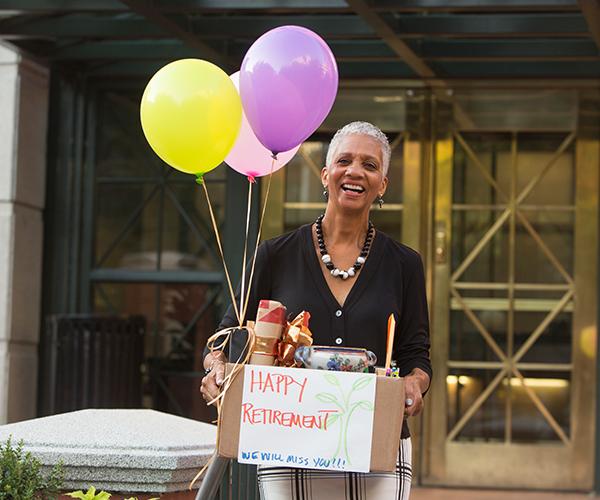 A person is standing outside carrying a box with items, inflated balloons and a piece of paper that reads happy retirement.