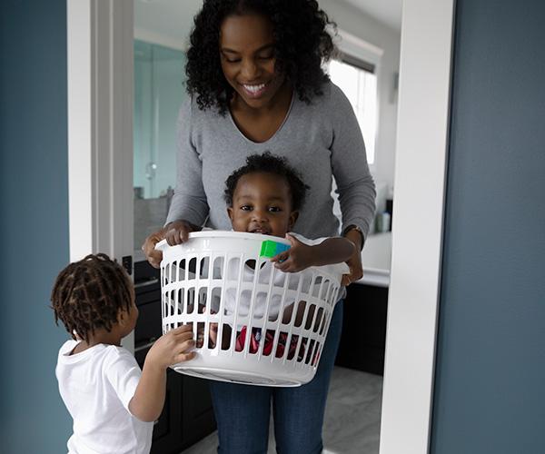 A person is holding a baby in a laundry basket in a doorway while another child stands facing towards the basket.