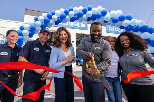 A group of people standing behind a red ribbon in front of a building while one person is cutting the ribbon with giant scissors.