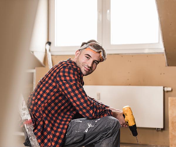 A person wearing safety goggles is crouched down and holding a drill in front of a wall with a window.