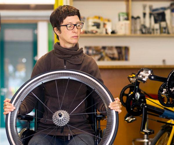 A person seated in a wheelchair holding a bicycle wheel looking at a bicycle pedal.