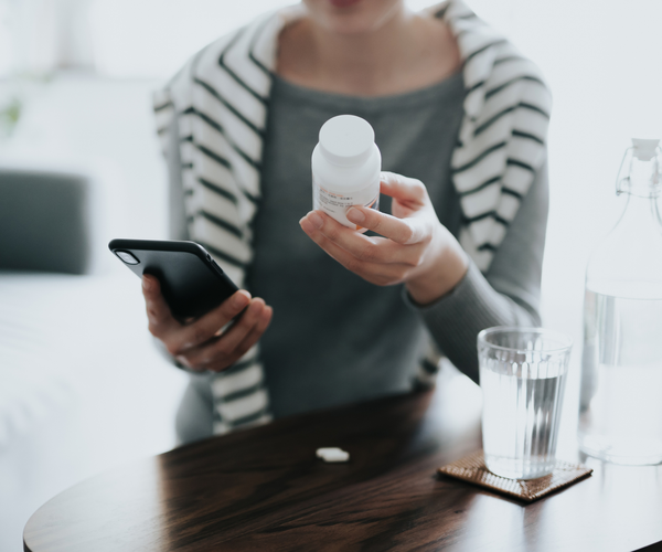 A person holding a medication bottle in one hand and a smartphone in the other, with a glass of water on the table in front of her.