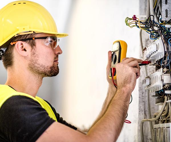 A person wearing a yellow safety helmet is holding a voltage tester up to an electrical panel.