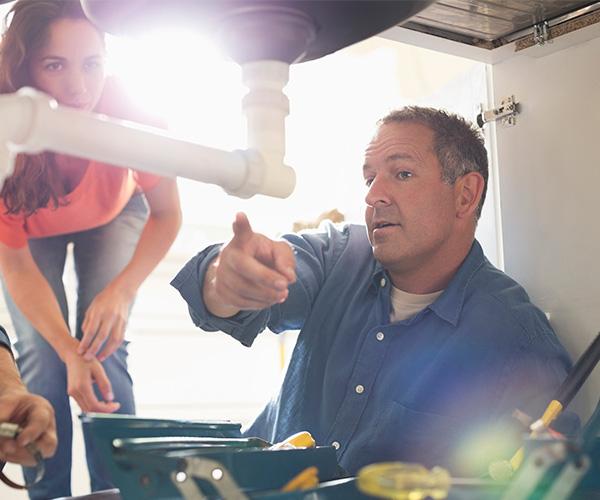 Two people are looking into an open cabinet under a sink. One of them is pointing at a pipe under the sink drain.