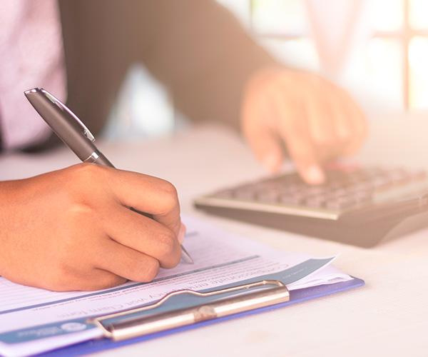 A person is seated at a desk writing on a piece of paper attached to a clipboard while pressing keys on a calculator.