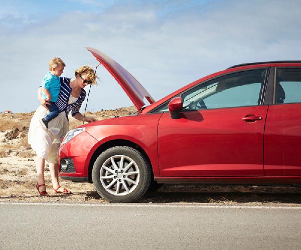 A person holding a child standing in front of a red car parked on the side of the road with the hood open.