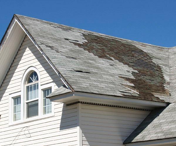 The roof of a white house with a section of missing and damaged gray shingles.