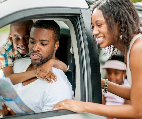 An adult and two children are seated in a vehicle looking at a map while another adult is standing outside of the vehicle looking at the same map.