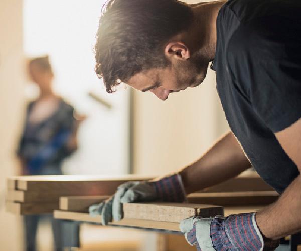 A person wearing gloves is holding two pieces of wood together. A person in the background is holding a paint roller.