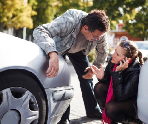 One person is bent at the waist to talk to another person who is crouched in front of a gray sedan talking on a cell phone.