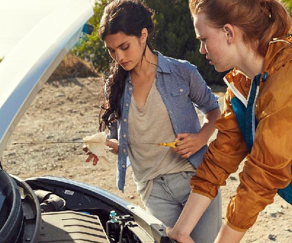 Two people standing in front of a car with the hood open looking inside. One of them is holding an oil dipstick.