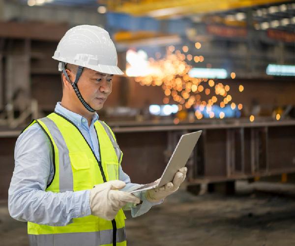 A person wearing a green vest and white safety helmet holding a laptop in a warehouse.