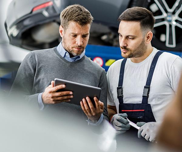 The man on the left, in a grey sweater, is showing a tablet to the man on the right. The man on the right is holding a wrench and wearing a black apron.