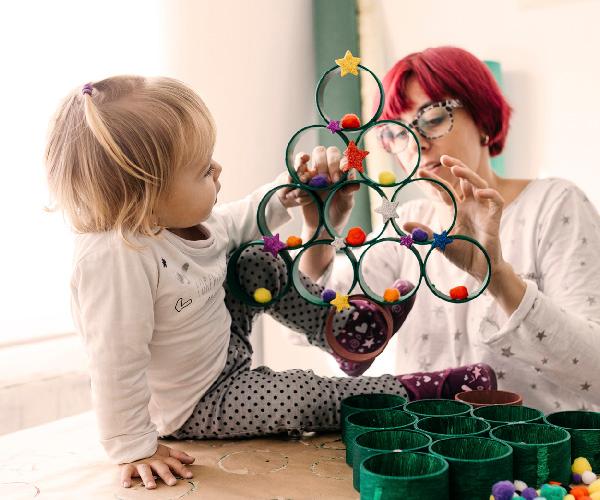 A child is sitting on a table holding an arrangement of green circles of paper to resemble a tree with an adult helping them.