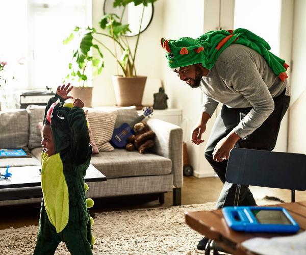 A child in a dinosaur costume is standing in a living room while an adult stands behind them wearing a dinosaur costume on their head.