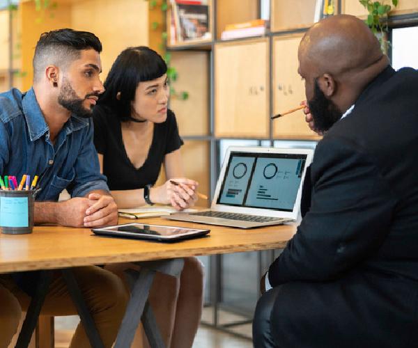 Two people seated at a table are looking at a person seated across from them who is pointing towards a laptop with graphs on it.