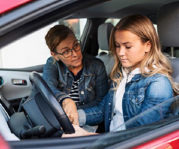 A young driver is seated in the driver seat of a red vehicle while an older passenger is instructing them.