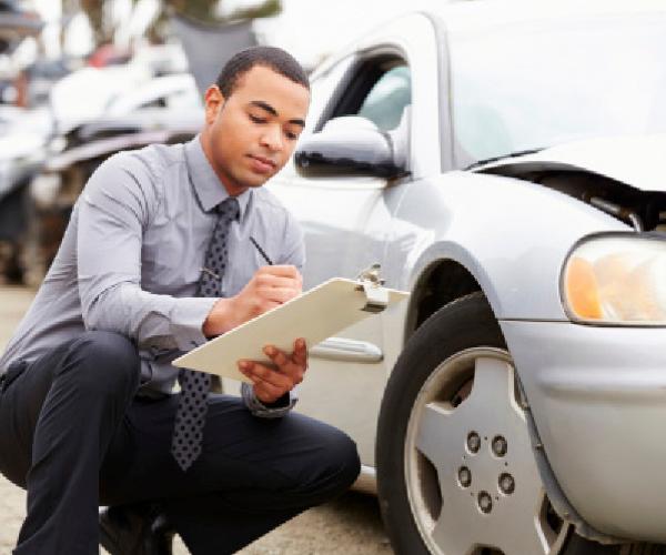 A person with a clipboard and pen is crouched next to a silver sedan with damage to the hood.