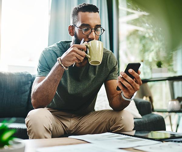 An individual sitting on a couch drinking coffee while looking at a phone.