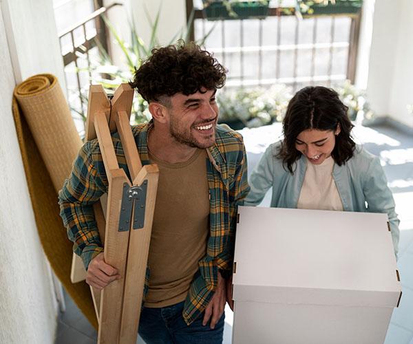 Two individuals walking together. One is carrying a step ladder and the other is carrying a cardboard box.