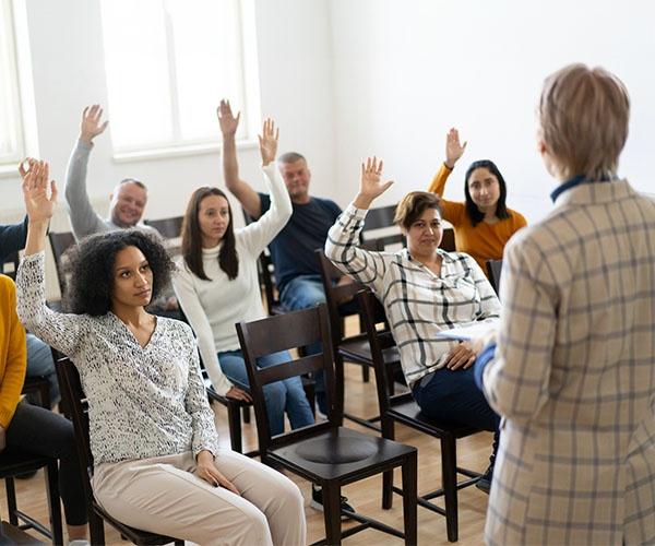 Six people seated in a room with their hands raised. One person is standing at the front of the room facing those six people.