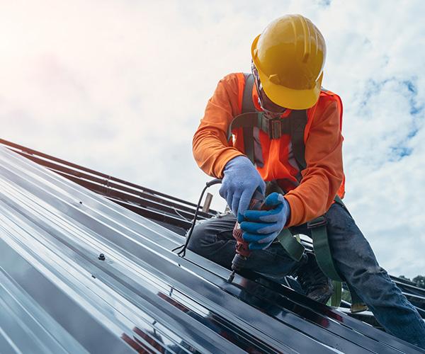 A person on a roof who is wearing a construction hat and using a tool to install shingles in place.