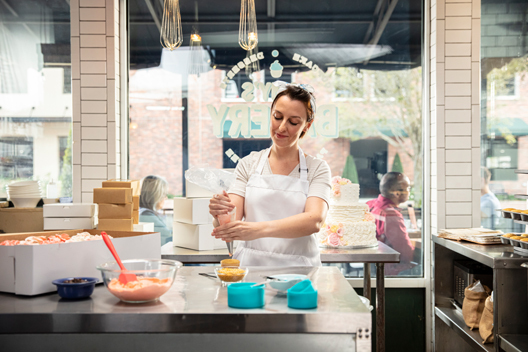 A person standing in a bakery holding a piping bag, putting frosting on baked goods.