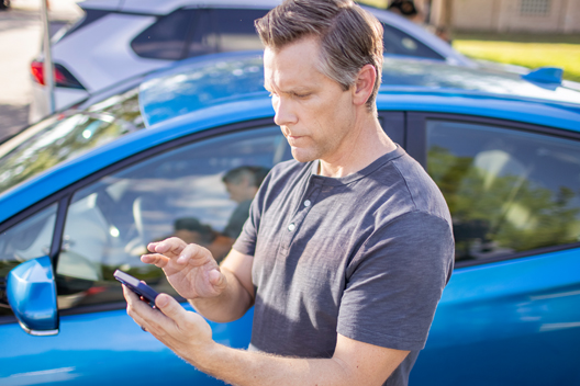 A person with short brown and grey hair standing in front of a car, holding a phone.