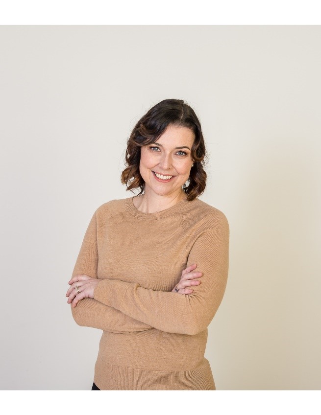 A lady with brown hair standing with her arms folded wearing a light brown sweater