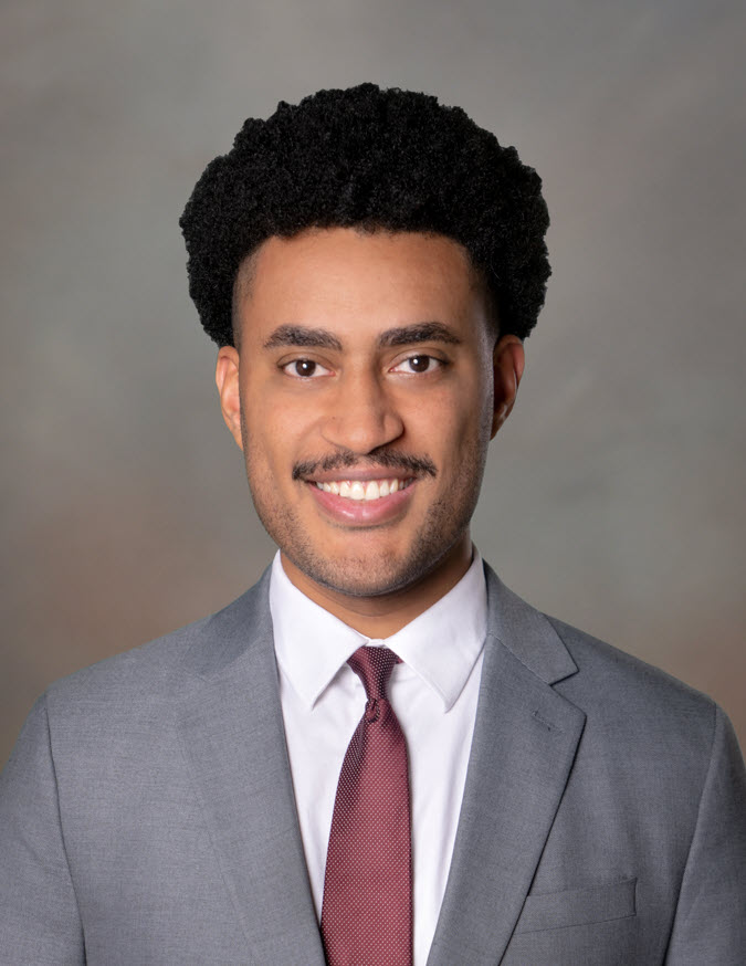 A person with short black hair wearing a white shirt with a grey blazer and a burgundy tie, against a brown background.