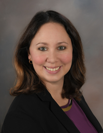 A person with medium brown hair wearing a purple shirt with a black blazer, against a brown background.