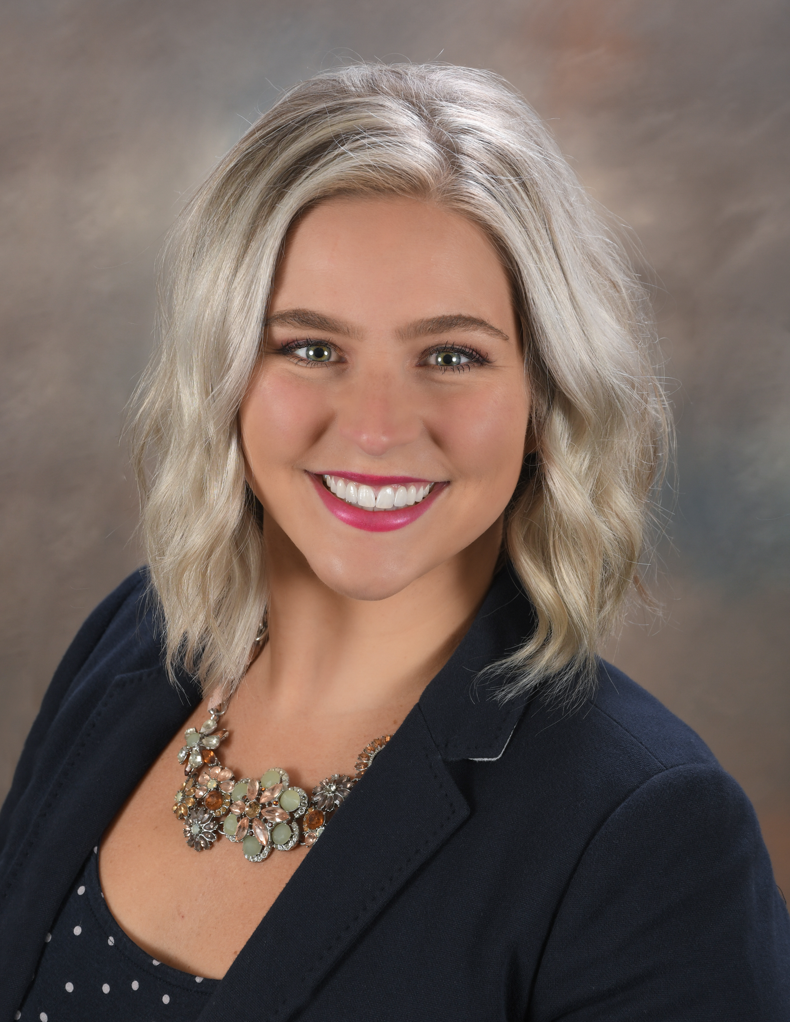 A person with medium blonde hair wearing a black shirt with a black polka dot blazer, against a brown background.