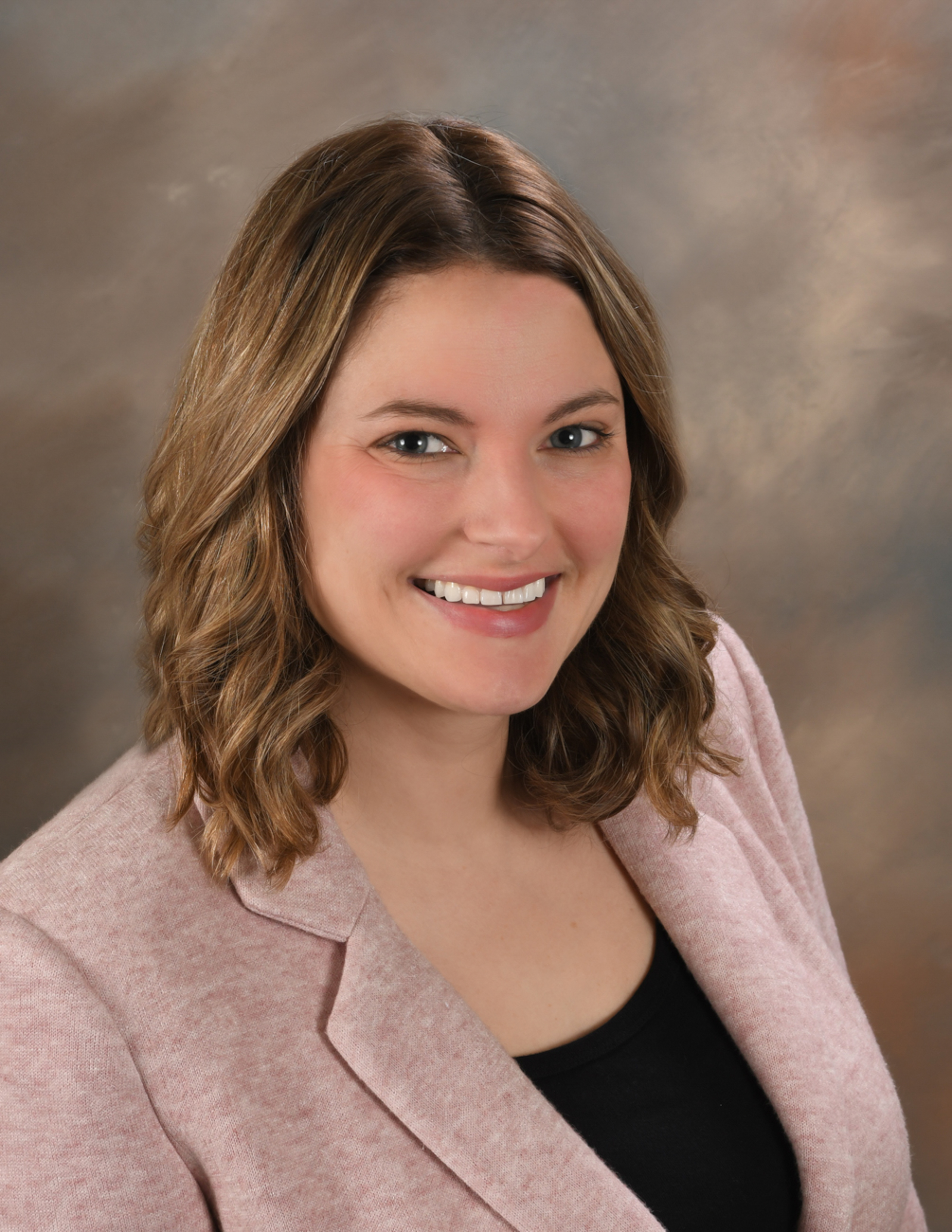 A person with medium brown hair wearing a black shirt with a pink blazer, against a brown background.