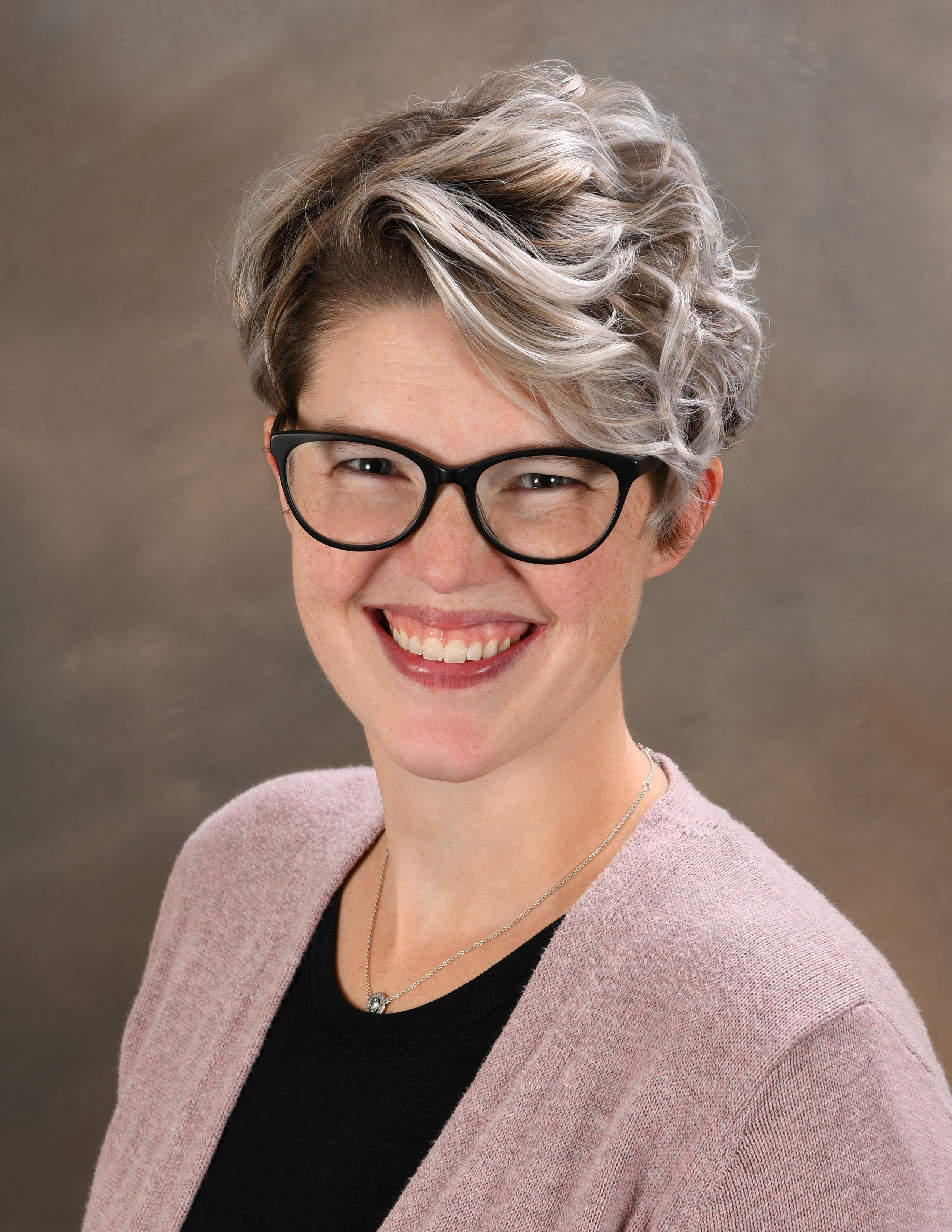 A person with short blonde hair wearing a black shirt with a pink blazer, against a brown background.