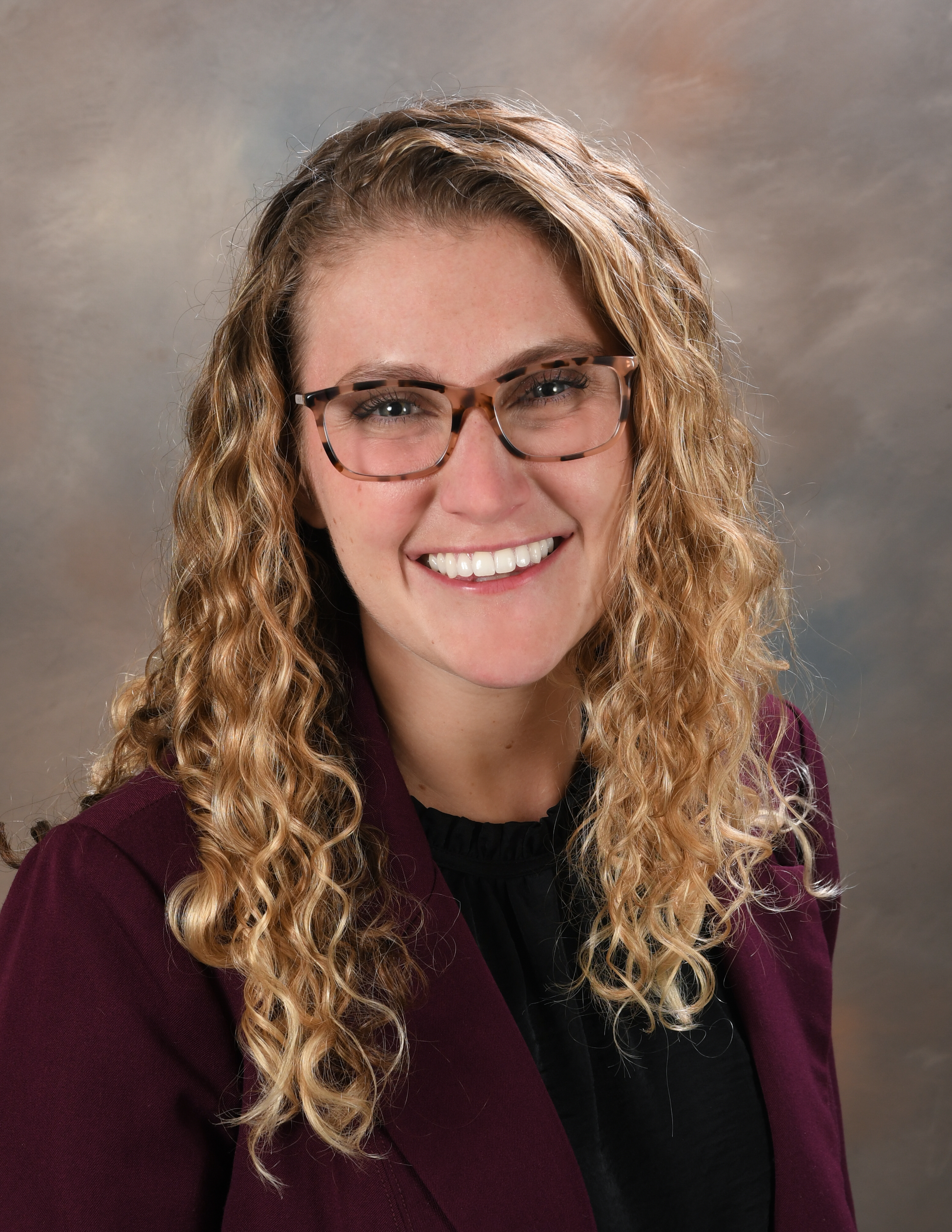 A person with long blonde hair wearing a black shirt with a burgundy blazer, against a brown background.