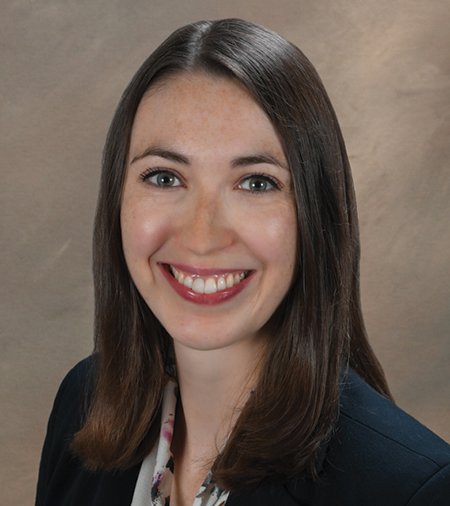 A person with medium brown hair wearing a white shirt with a black blazer, against a brown background.