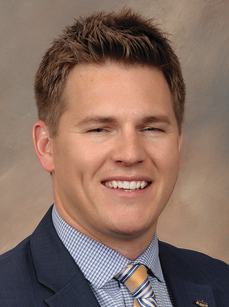 A person with short brown hair wearing a blue plaid shirt with a black blazer and a blue and yellow tie, against a brown background.