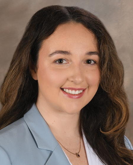 A person with long brown hair wearing a white shirt with a light blue blazer, against a brown background.