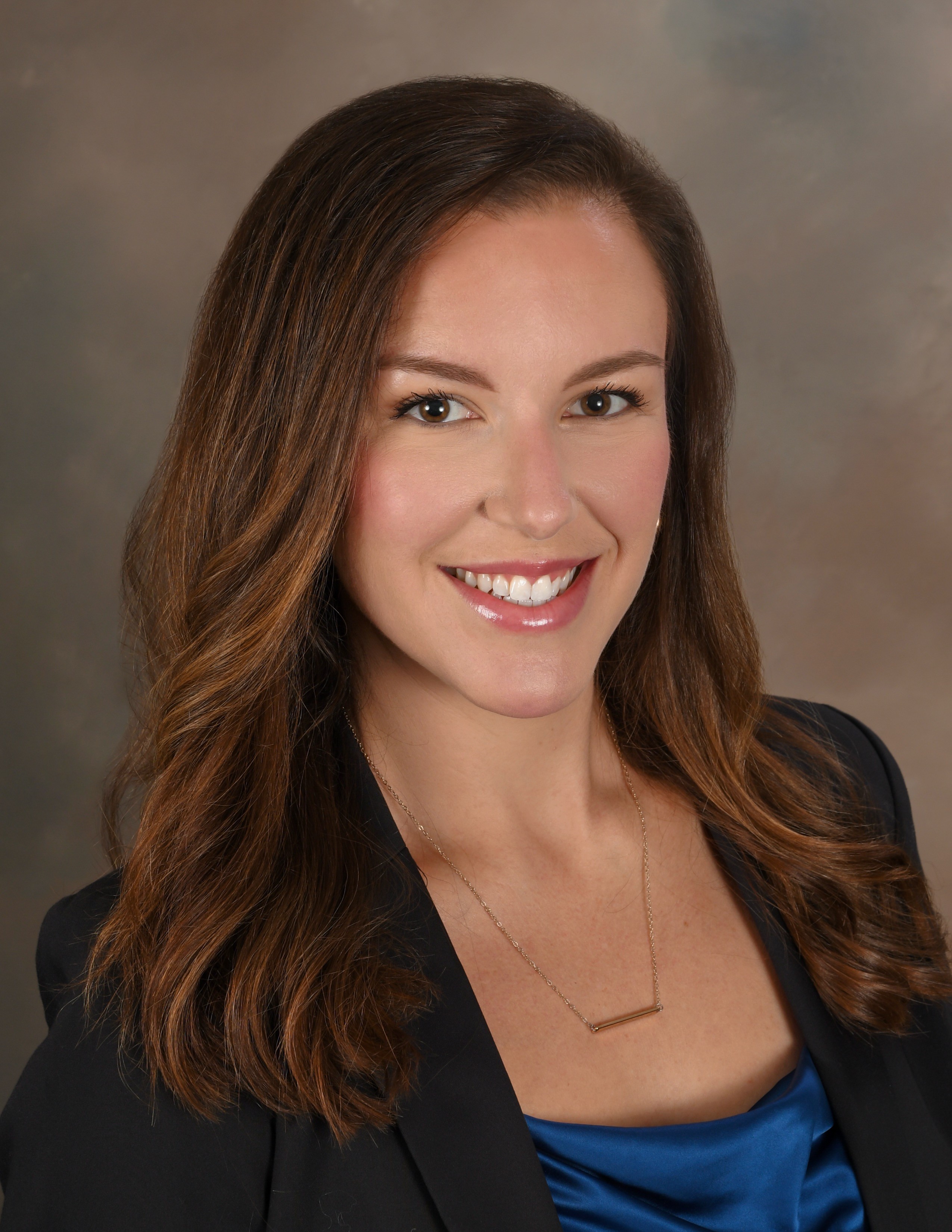A woman with dark brown hair wearing a blue shirt with a black blazer, against a grey background.