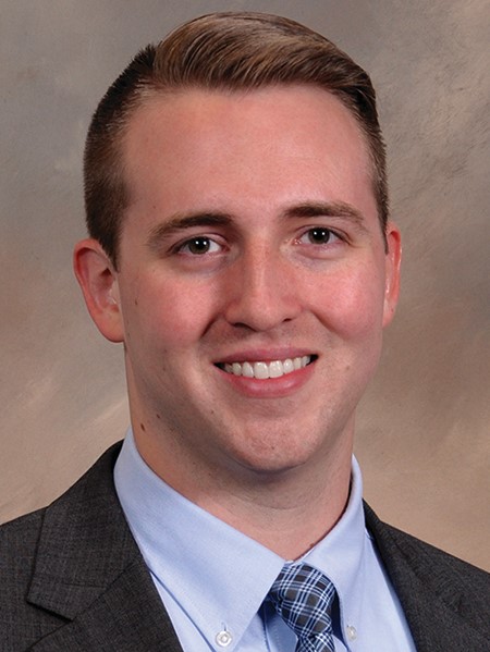 A person with short brown hair wearing a light blue shirt with a dark grey blazer against a brown background.